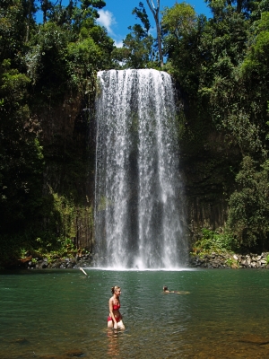 A woman stands by Paronella Park’s scenic waterfall amid vibrant rainforest, whilst another person swims in the refreshing natural pool.