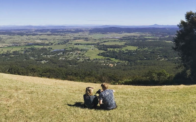 Two people relax on a lush grassy hill overlooking scenic Gold Coast valleys, envisioning a luxurious Ultimate Gourmet Tasting Experience.