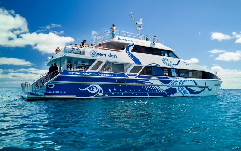 The Aquaquest Outer Reef Snorkel Special catamaran glides on crystal-clear, tranquil water beneath a radiant blue, sunlit sky.