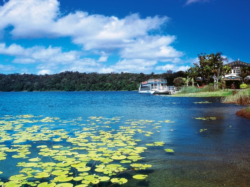 Serene northern lake with vibrant lily pads, a quaint boat by Paronella Park’s scenic shore beneath a picturesque blue, cloudy sky.