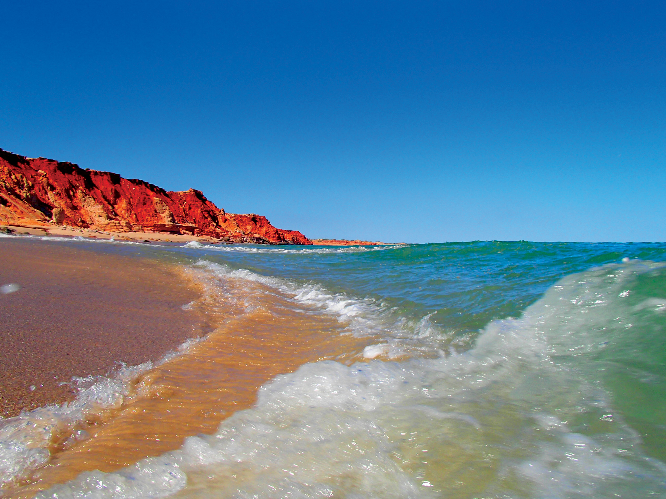 Powerful waves crash onto a pristine sandy beach bordered by striking red rocky cliffs on the 10 Day Broome to Perth Adventure.
