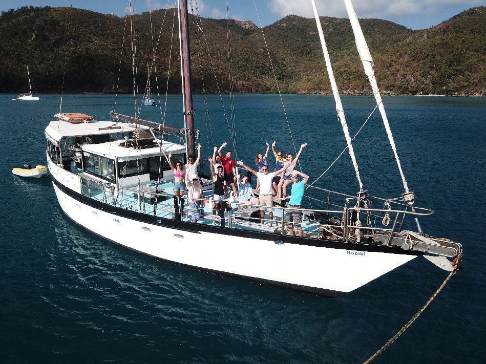 Friends enjoying the Habibi 2 Day 1 Night Whitsunday Sailing adventure, anchored in turquoise waters beside lush green hills.