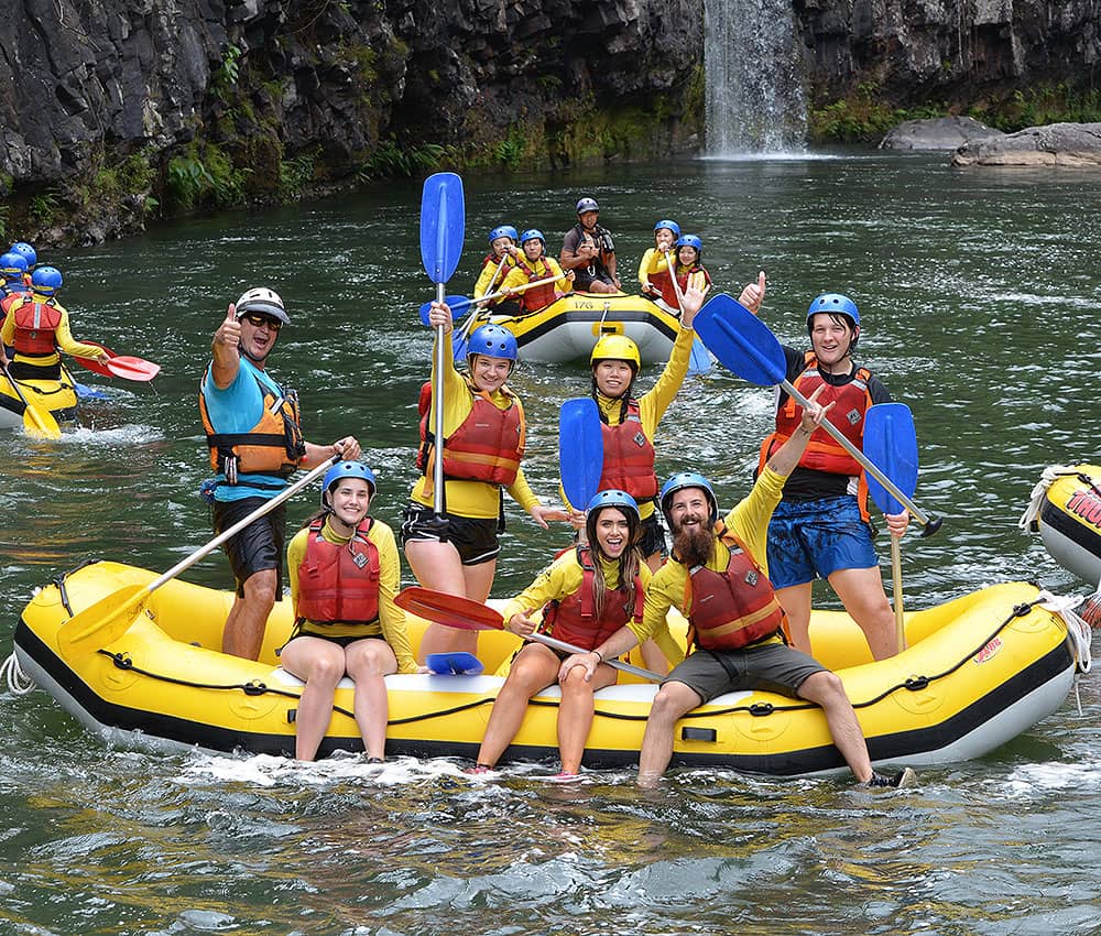 Thrill-seekers white water raft Barron River with Raging Thunder, smiling by stunning waterfall on a half-day adventure tour.