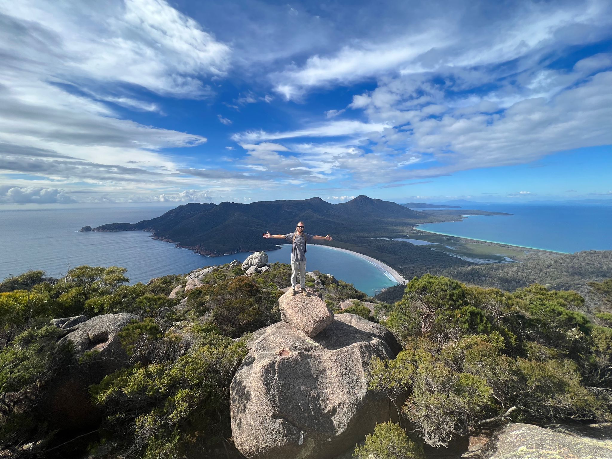 Traveller standing atop a rugged coastal rock, arms wide, enjoying panoramic sea views on the 6 Day Tasmania Explorer Tour adventure.