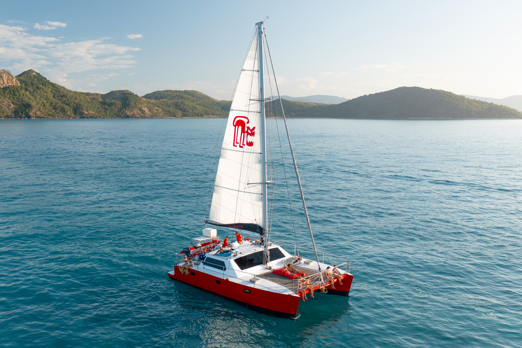 Red and white Tongarra-style catamaran sailboat cruising tranquil blue Whitsundays waters, lush green hills in scenic backdrop.