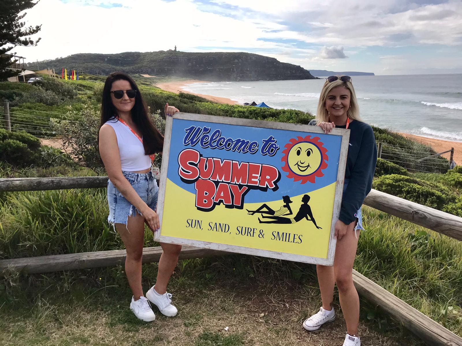 Two smiling women in summer attire hold a Welcome to Summer Bay sign, ideal for your 1 Day Home Away Location Tour experience.