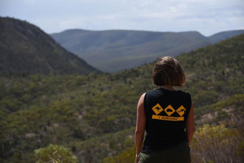 Traveller in a black "I Survived" shirt overlooks lush green hills during the 10-Day Adelaide to Perth Adventure Tour with Untamed Escapes.