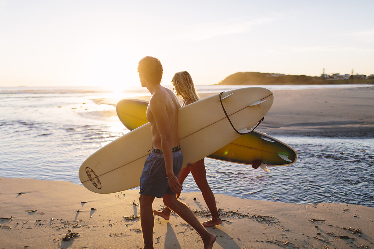 Two travellers with surfboards stroll a golden sandy beach at sunset, kicking off their 10 Day Oz Intro Sydney to Brisbane adventure tour.