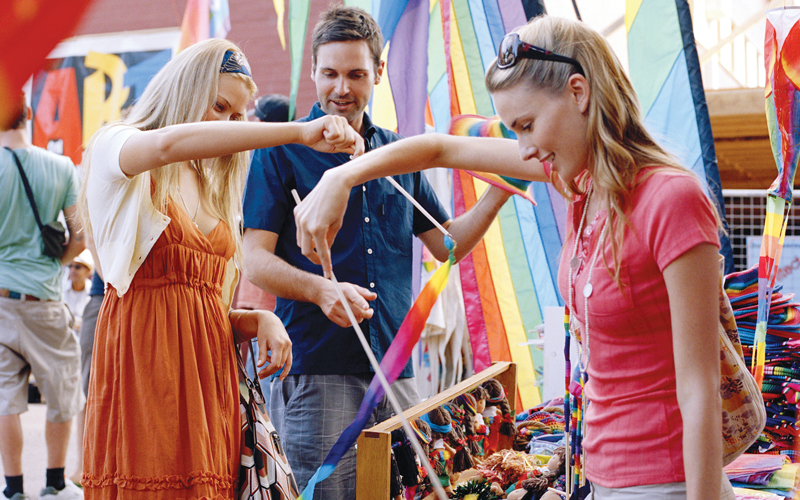 Three friends browsing vibrant handmade crafts and smiling at the lively Chill Out At Byron Bay outdoor market festival.