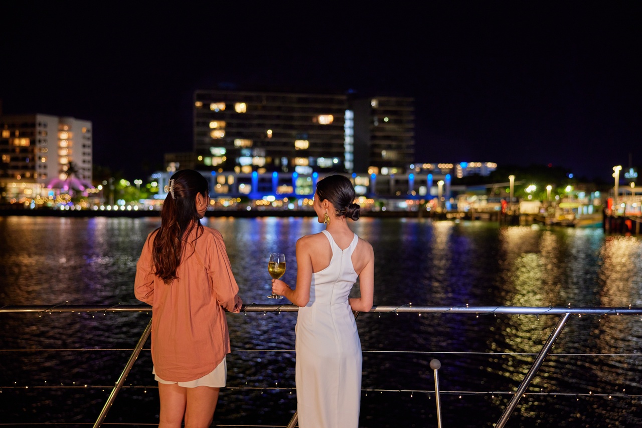 Two women enjoy a night cruise on the Spirit Of Cairns, vibrant harbour lights reflecting as one sips wine onboard the dinner boat.