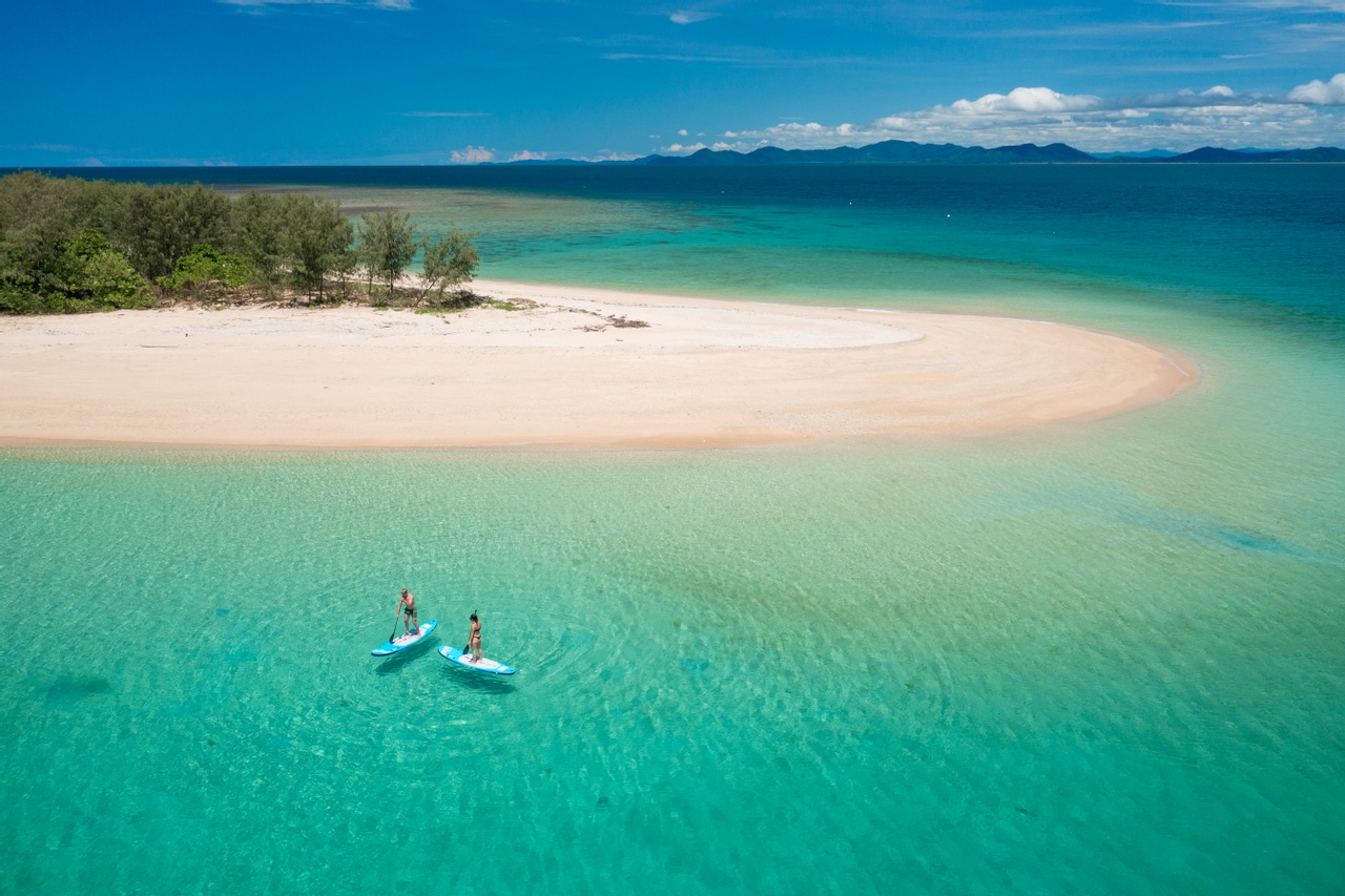 Two adventurers paddleboard in crystal-clear turquoise waters by a pristine sandy island on a Frankland Island Express Tour.