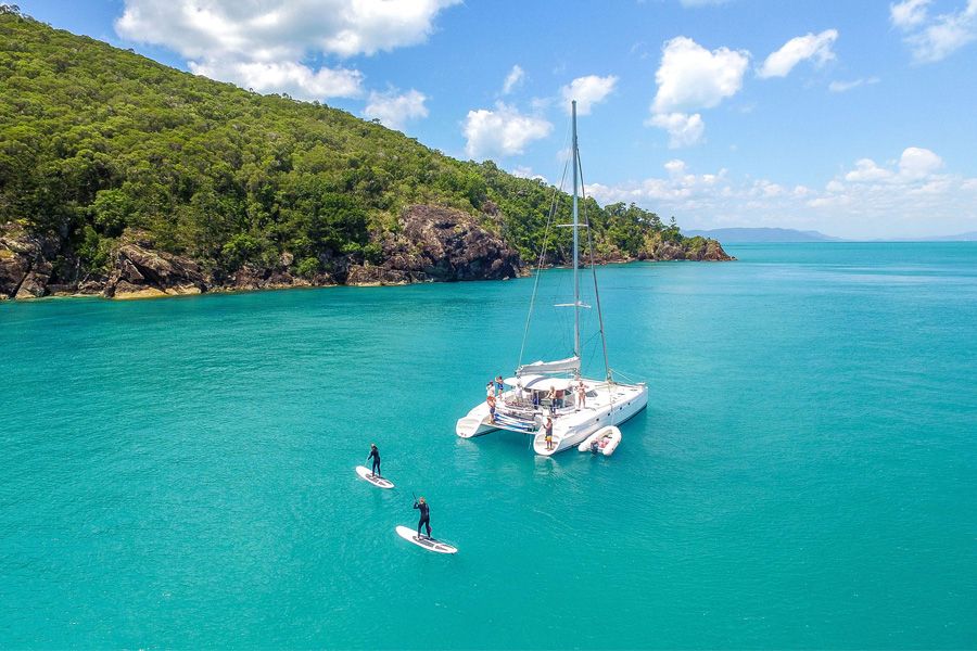 Whitsunday catamaran anchored in crystal-clear turquoise water, paddleboarders enjoying sunshine and blue skies, perfect sailing holiday.