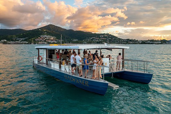 Experience a Sundowner Sunset Cruise on a floating boat bar as guests relax, with scenic mountains and shoreline views at golden sundown.