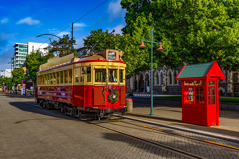 Vintage red tram and classic phone box on a sunlit Christchurch street, framed by leafy trees and iconic historic architecture.