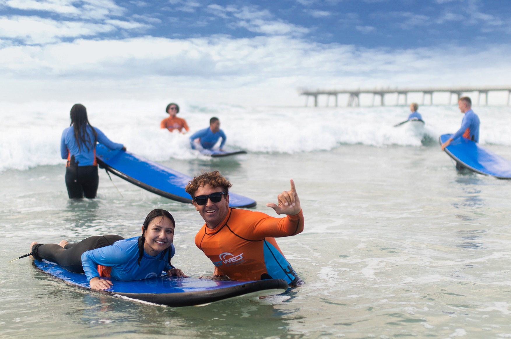 Teens and adults, 13+, grinning on a surfboard by the pier during a 2-hour beginner surf lesson—perfect for first-time surfers.