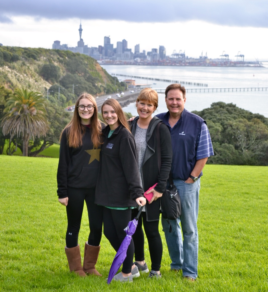Group of friends smiling on a grassy hill overlooking Auckland skyline and harbour, capturing iconic city highlights and vibrant atmosphere.