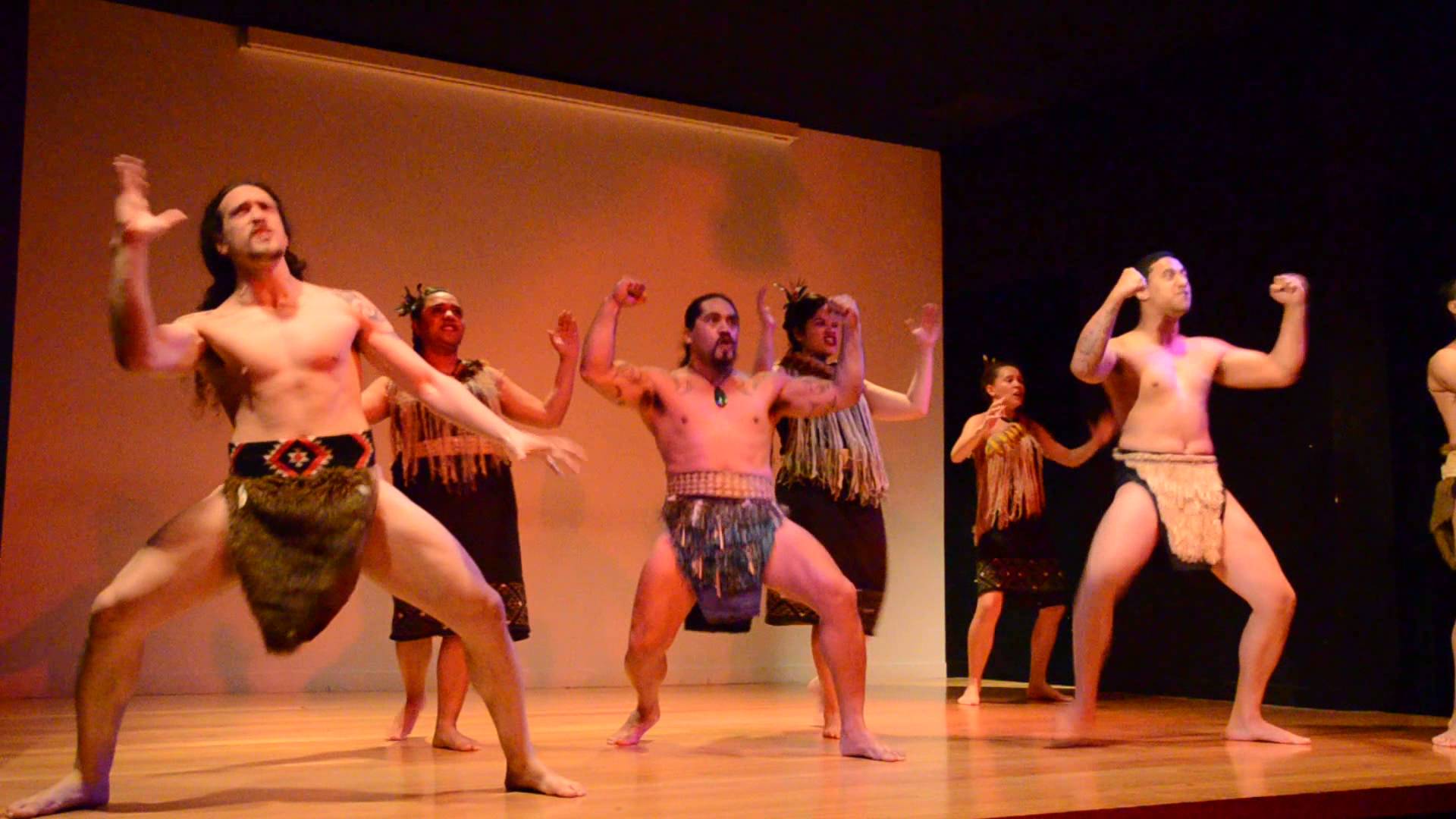 Traditional Māori performers in Auckland present an authentic haka dance in vibrant attire on a wooden stage during a Māori Experience.
