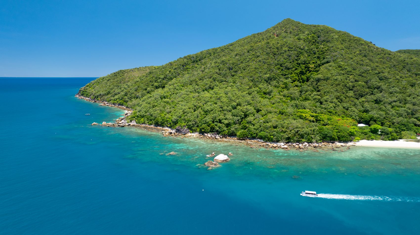 A boat glides by Fitzroy Island on the Island Value Package, surrounded by tropical greenery and sparkling blue waters in bright sunlight.