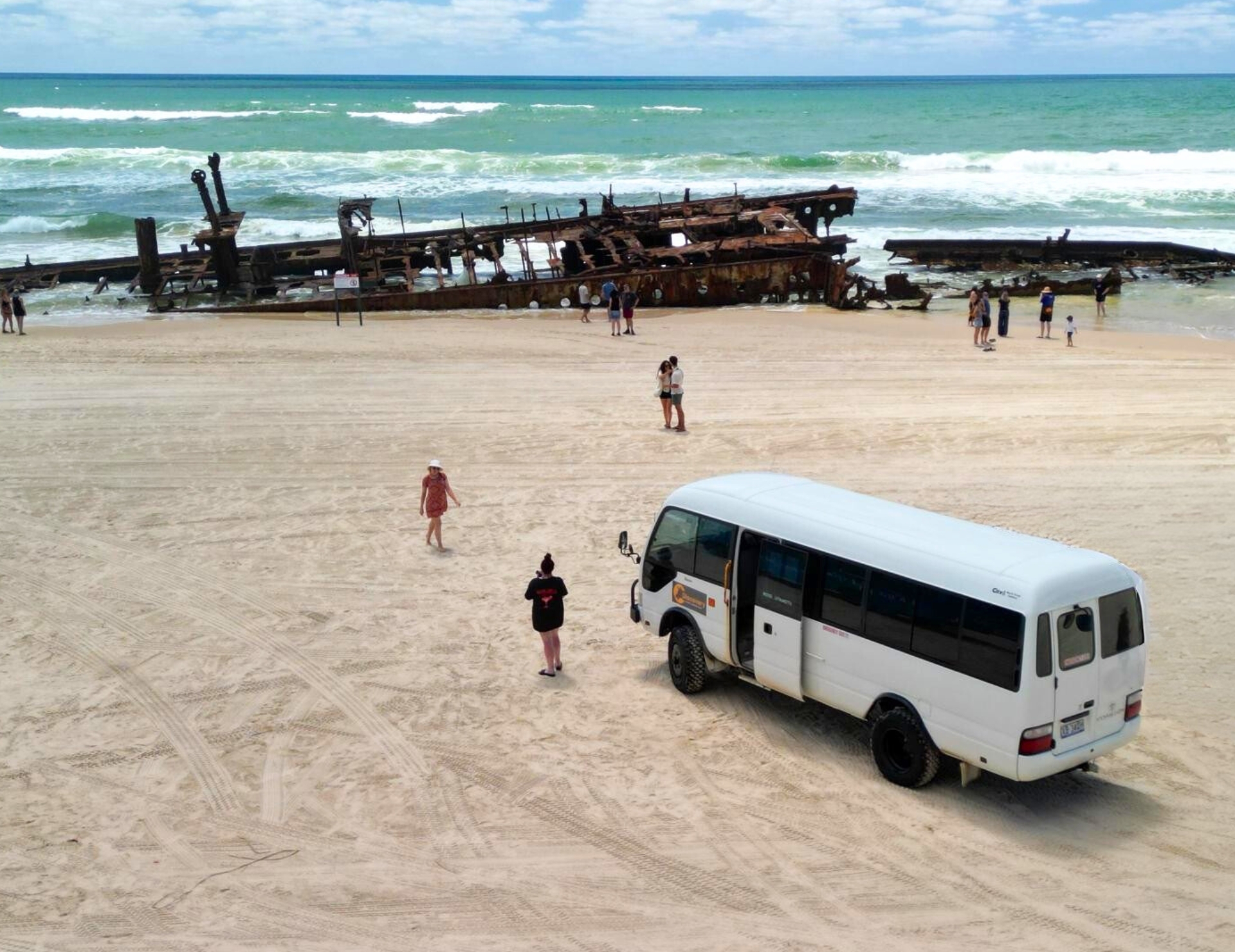 Fraser Island 2 Day Tour van, tourists on sandy beach beside iconic rusted shipwreck, scenic ocean view, top travel experience.