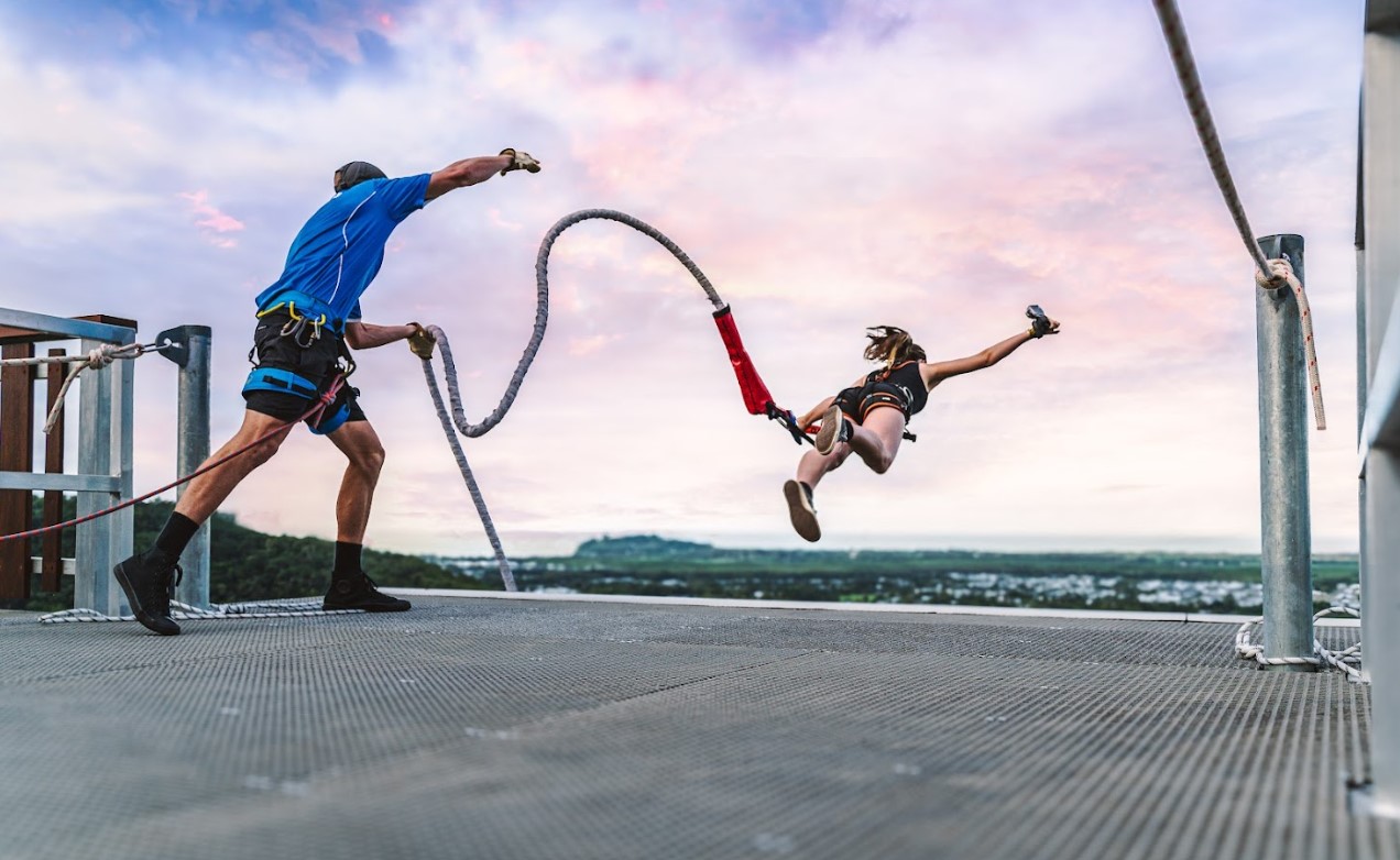 Thrill-seeker leaps for a Skypark Roof Jump at sunset as safety instructor fastens rope, highlighting extreme adventure sports experience.