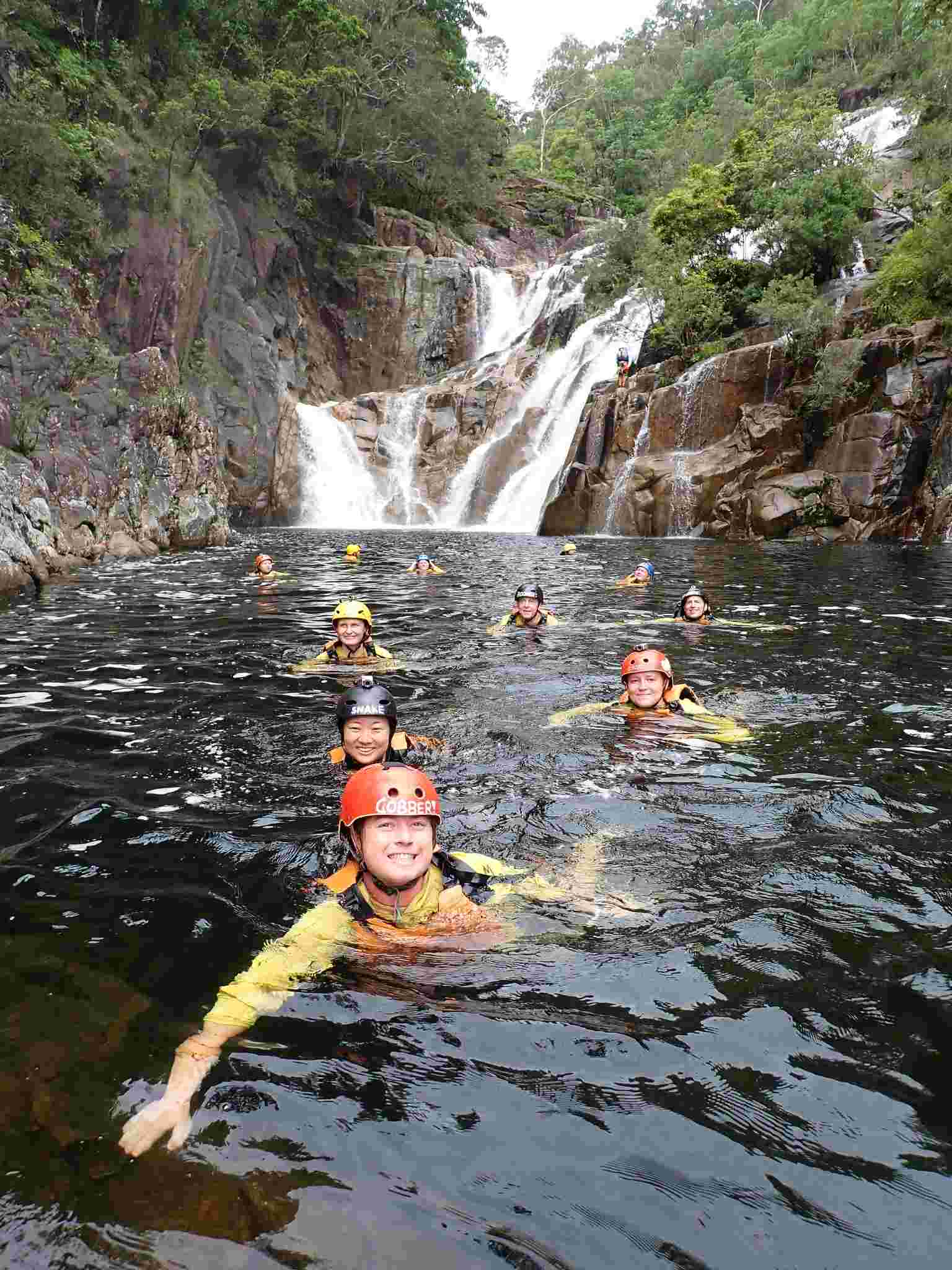 Adventurers in helmets swim through a rocky river beside a stunning waterfall on the Behana Gorge Canyoning Tour in Queensland.