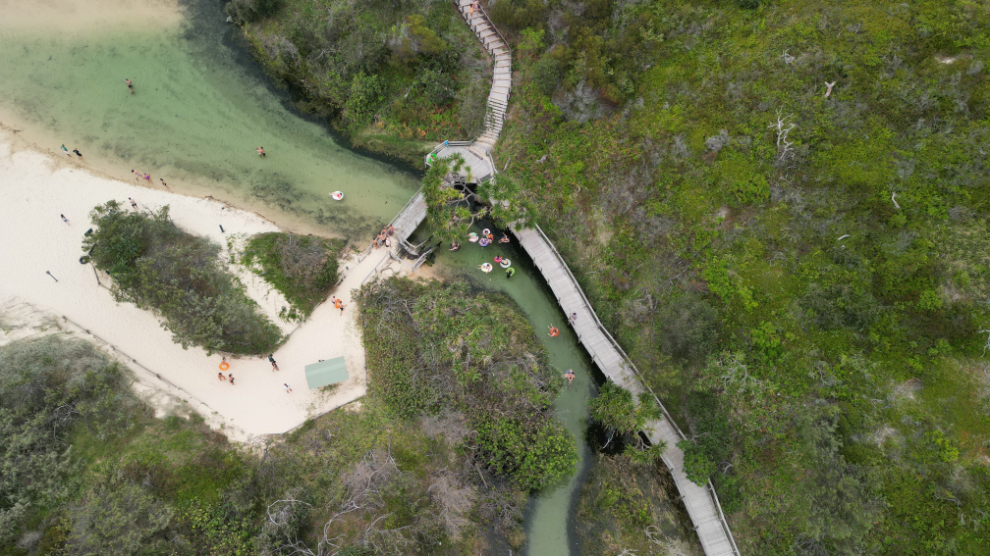 Stunning aerial view of Kgari 2 Day Tour’s winding boardwalk crossing a stream, guiding visitors to Noosa’s pristine sandy beach.