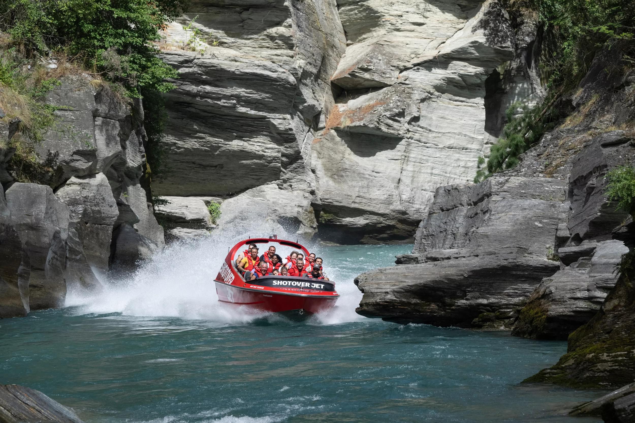 A bright red Shotover Jet boat races through a scenic rocky canyon over crystal-clear water, splashing mist as excited riders hold on.