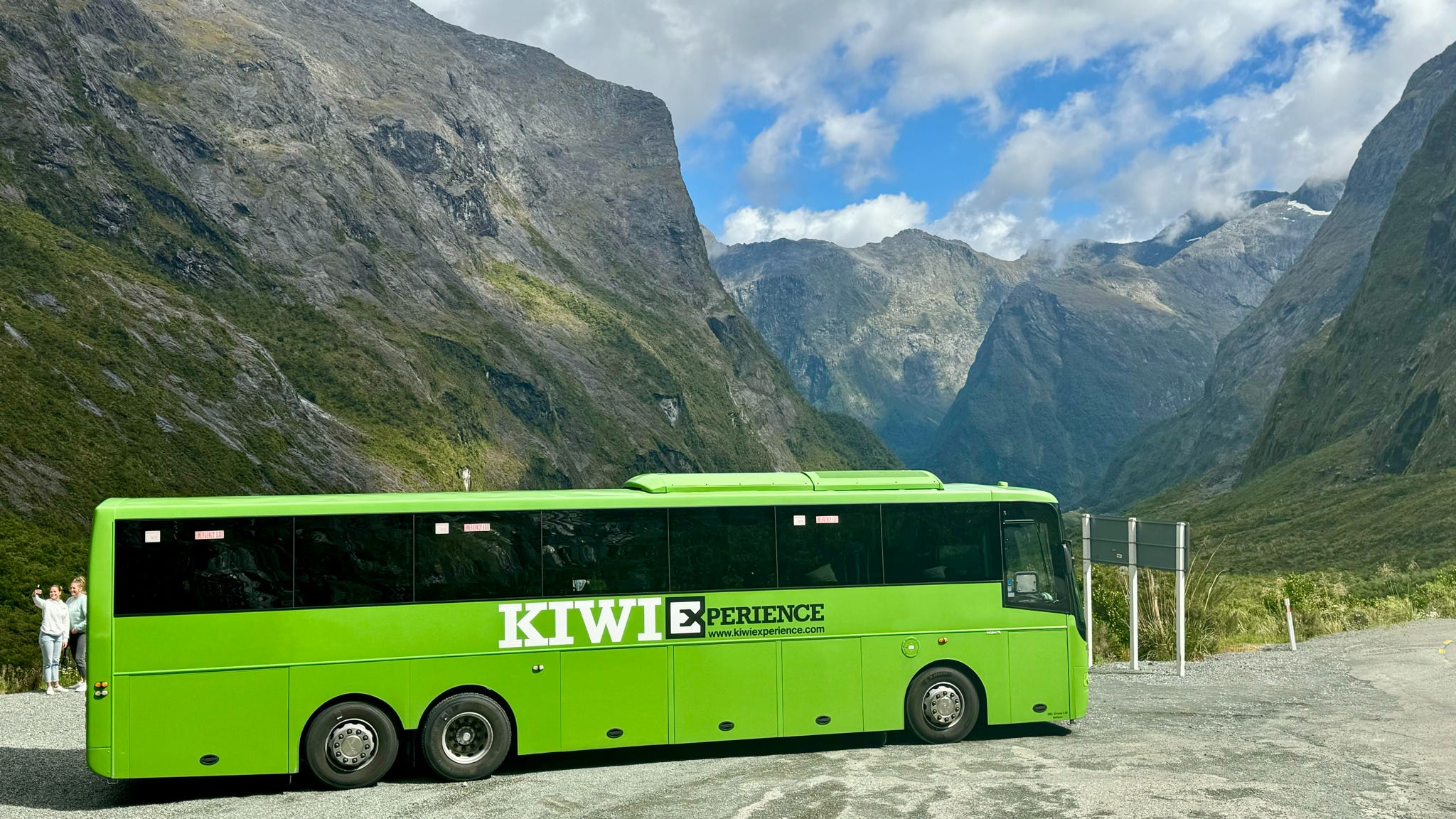 Large green Kiwi Experience bus with the background of Milford sound