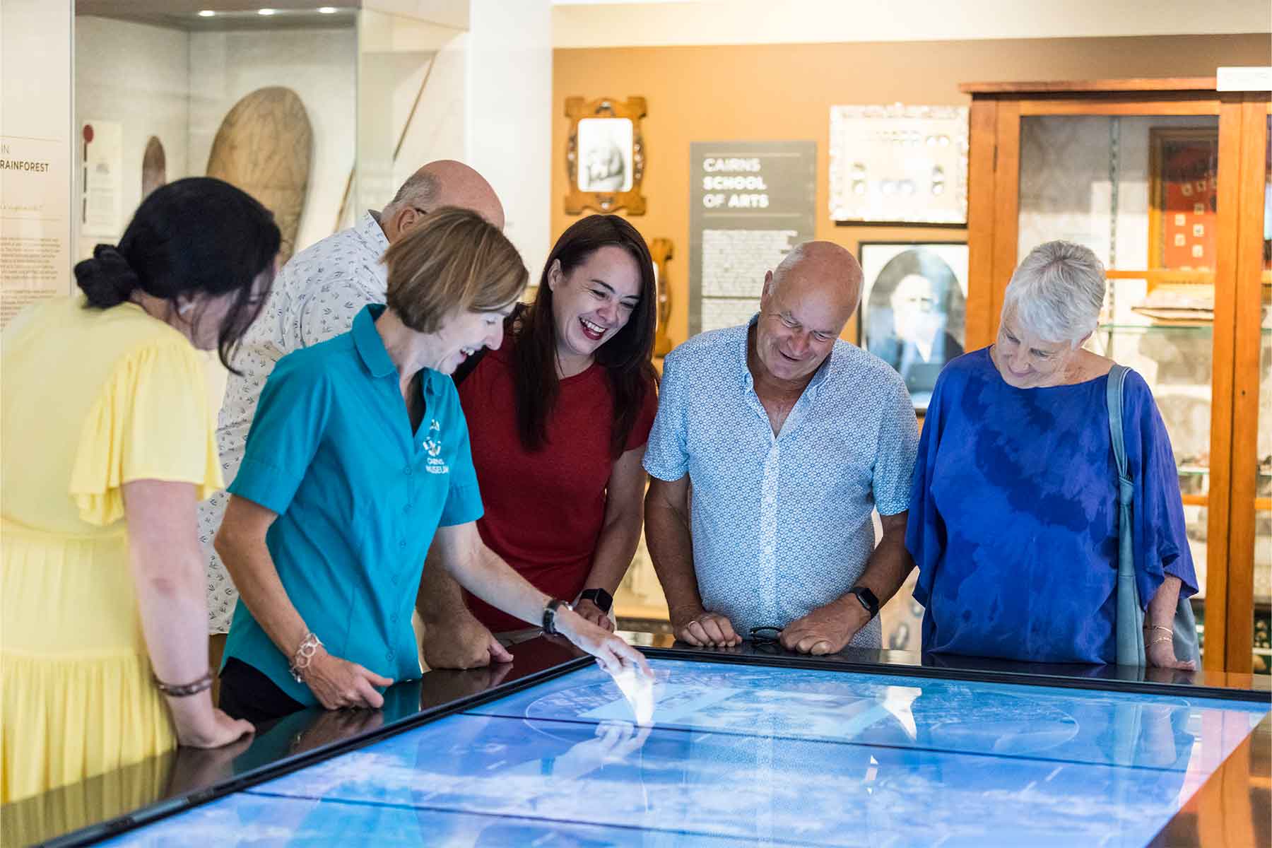 Six adults gather around an interactive touchscreen display during a CSHD Tour in a brightly lit Cairns City museum.