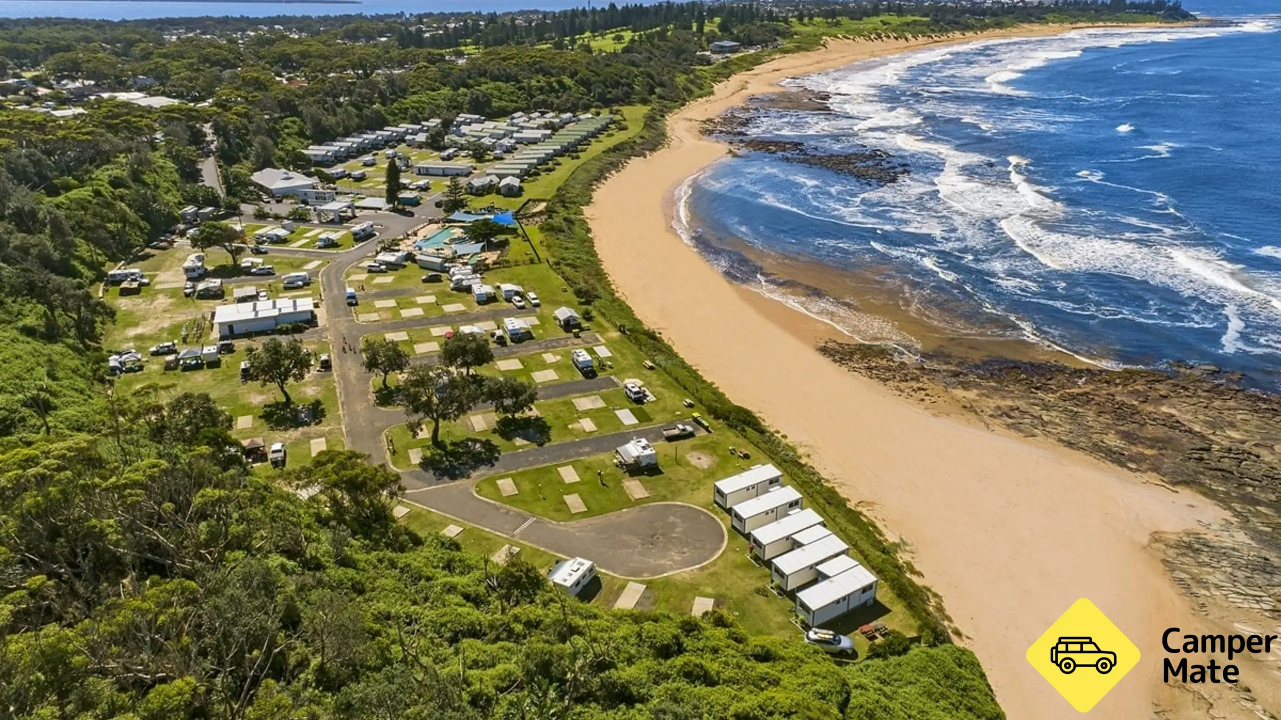 Ocean coast on the right with blue water and yellow sand. The grass behind the sand holds a busy caravan park with plenty of sites overlooking the ocean